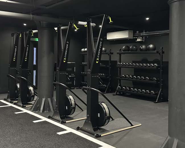 The image shows a modern gym interior featuring rowing machines and a rack of black weights on a dark, carpeted floor.