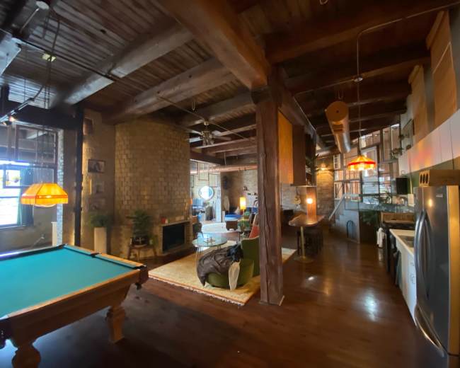 The image shows an open-concept loft with a billiard table in the foreground and various seating areas, including a couch and armchairs, along with exposed wooden beams and brick walls.
