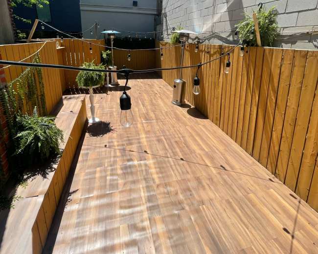 The image shows a wooden outdoor space with hanging lights and plants against a backdrop of a brick and gray wall, under a clear blue sky.