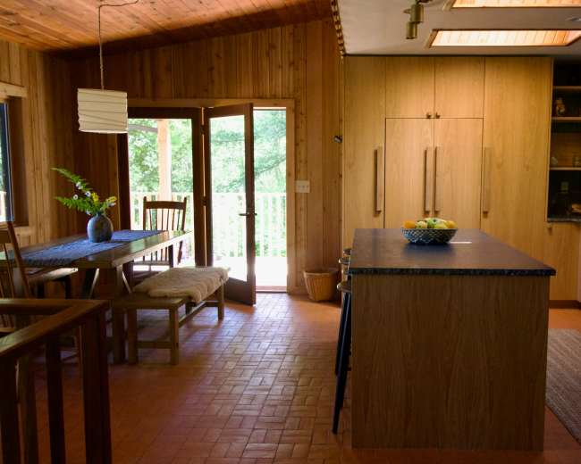 The image shows a warm wood-finished kitchen and dining area with a large island, a wooden dining table, and open doors leading to a deck.