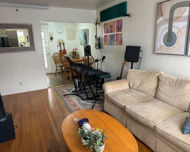 A living room with a beige couch, a wooden coffee table, and a keyboard set up near a dining area that has wooden chairs and a table.
