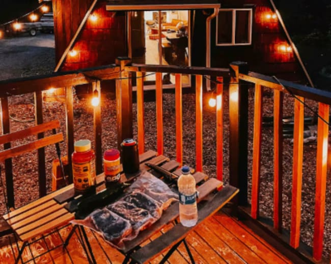 A wooden A-frame cabin is illuminated by string lights, with a table set for a barbecue on the deck.