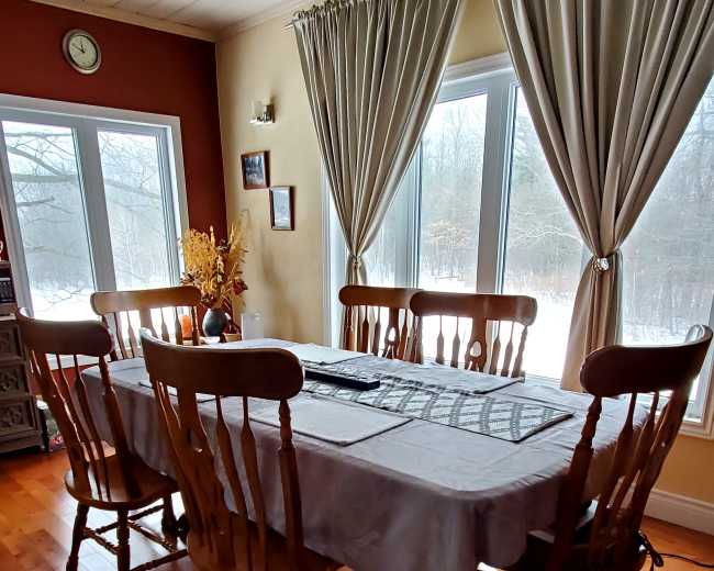 A large wooden dining table is set with a tablecloth in a well-lit room featuring large windows and wooden flooring.