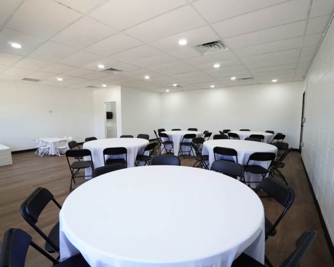 The image shows a spacious room with several round tables covered with white tablecloths and black chairs arranged around them.