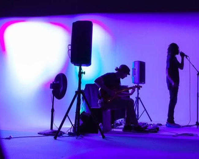 A musician stands at a microphone while another plays guitar in a dimly lit setting with purple lighting.