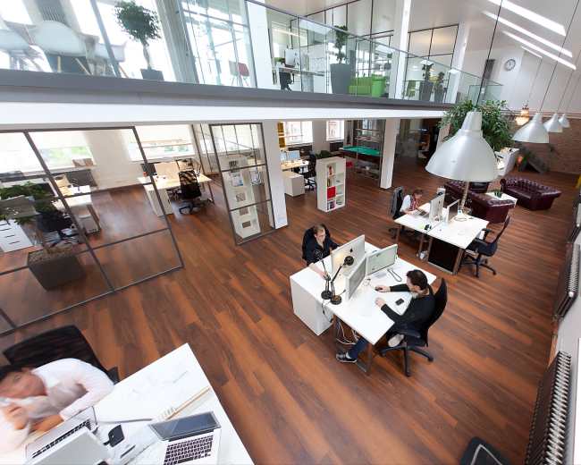 The image shows a modern office space with several workstations, large windows, and a mix of open and enclosed areas.