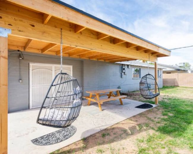 The image shows a backyard with a covered patio featuring two hanging chairs and a wooden picnic table on a concrete surface.