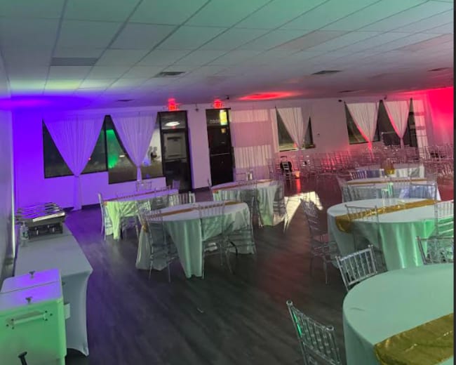 The image shows a spacious event venue with white tablecloths on round tables, decorated with gold accents, and illuminated by green and red lighting.