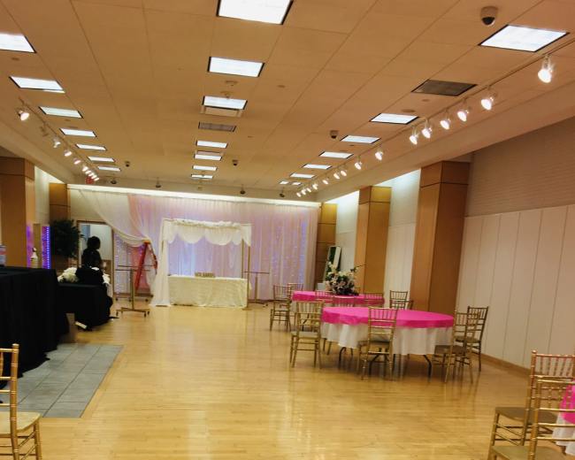 The image shows a spacious event hall with tables dressed in pink and gold chairs, set up for a celebration.