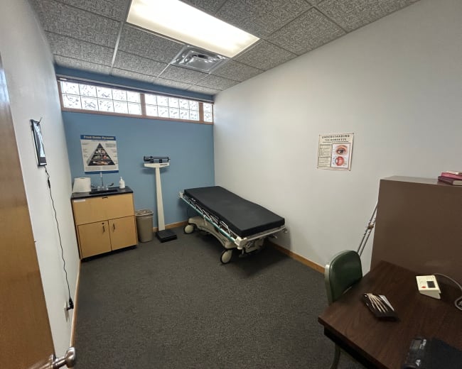 The image shows a small examination room featuring a medical examination table, a scale, a desk with a chair, and a waste bin.