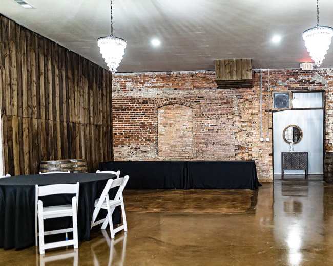 The image shows an empty room with exposed brick walls, a polished concrete floor, a table covered in a black cloth, and two chandeliers hanging from the ceiling.