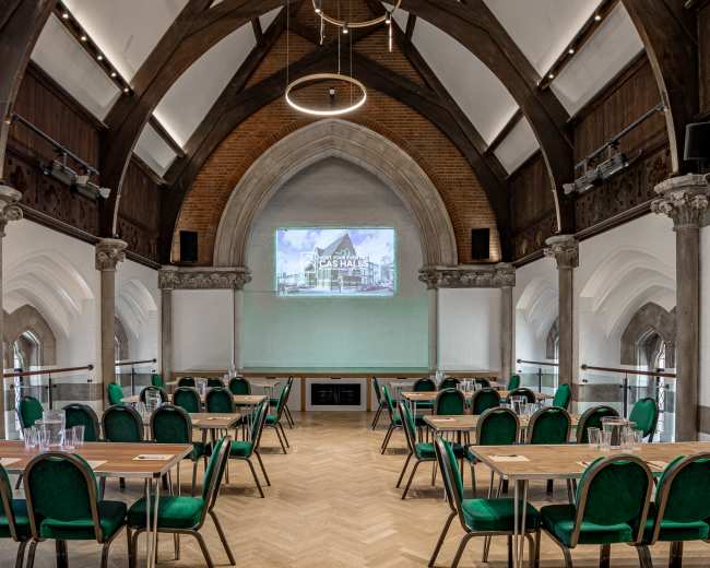The image shows a spacious meeting room with high ceilings, equipped with tables and green chairs arranged for a presentation, and a projector screen at the front.