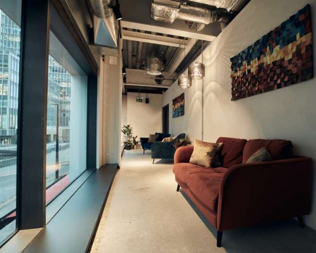 The image shows a modern interior space featuring a red couch, two blue chairs, and colorful wall art, with large windows providing natural light.