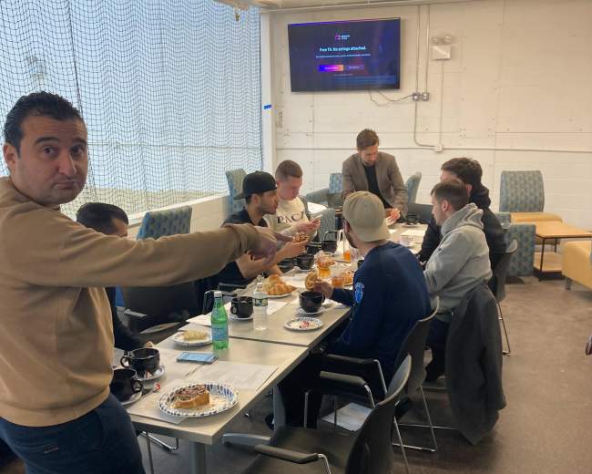 A group of people is sitting around a table filled with food, while one person is standing and gesturing towards the camera.