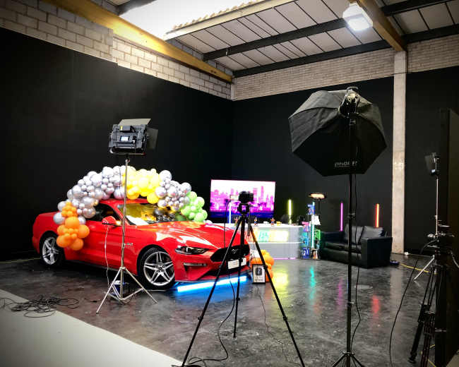 A red car is decorated with colorful balloons in a studio setting equipped with lighting and a television display.