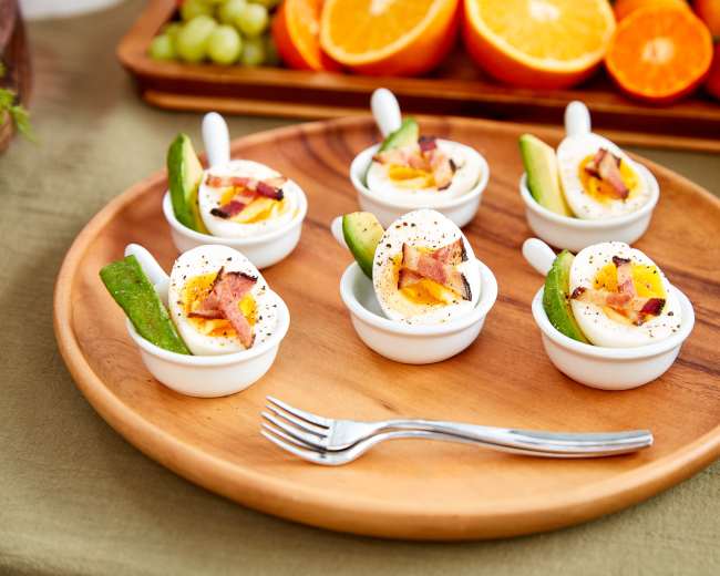 The image shows six servings of deviled eggs garnished with bacon and green pepper on a wooden plate, accompanied by a fork.