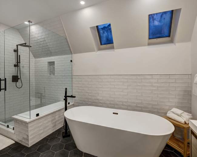 The image shows a modern bathroom featuring a freestanding bathtub, a glass shower enclosure, and angled skylight windows.