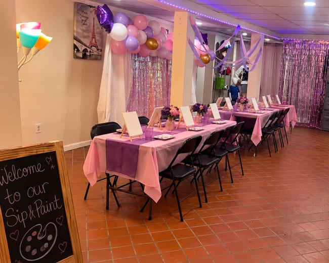 The image shows a decorated event space with rows of tables set up for a paint and sip party, featuring pink tablecloths, floral decorations, and colorful balloons.