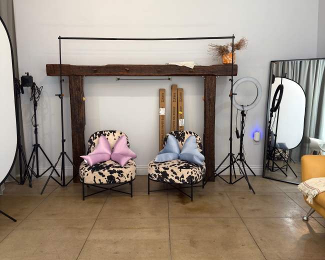 The image shows a minimalist studio setup with two patterned chairs, a large mirror, lighting equipment, and a wooden beam above.