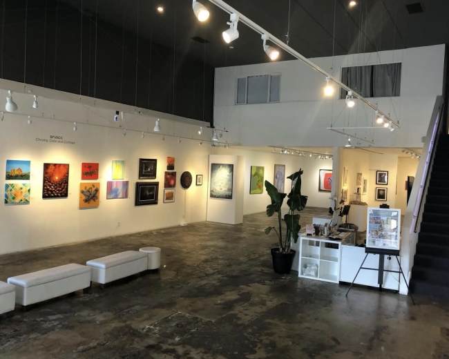 The image shows an art gallery with various paintings displayed on the white walls, a reception area with a desk, and stairs leading to an upper level.