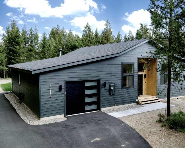 A modern grey house with a wooden entrance sits next to a paved driveway, surrounded by trees in a natural setting.