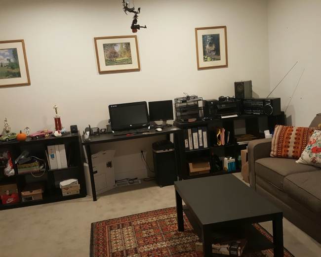 A living room features a sofa, a coffee table, a desk with two computer monitors, and shelves containing various items, including a trophy and framed pictures on the walls.