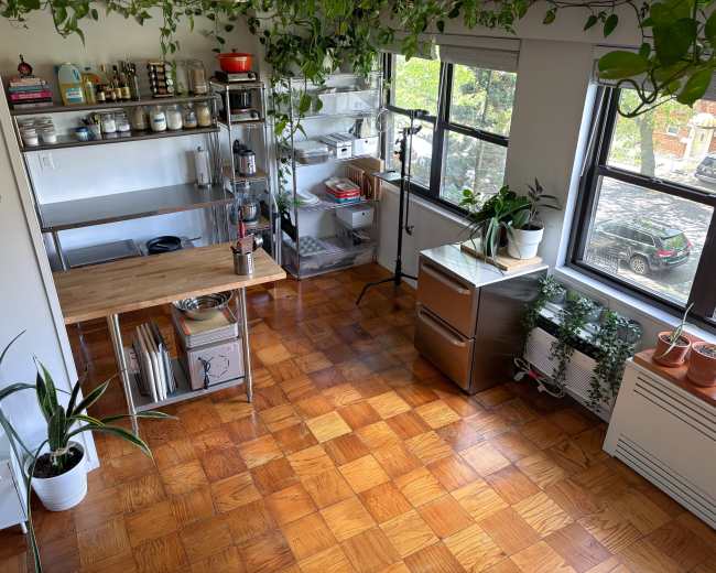 The image shows a bright kitchen with wooden flooring, filled with plants, metal shelving units, and a workshop-style table.