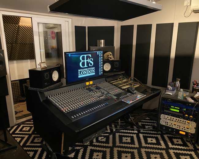 The image shows a recording studio with a large mixing console, studio monitors, and soundproofing panels on the walls.