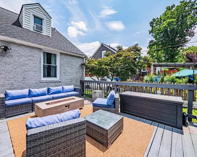 The image shows a spacious outdoor deck with modern wicker furniture arranged around a fire pit, surrounded by greenery and residential buildings.