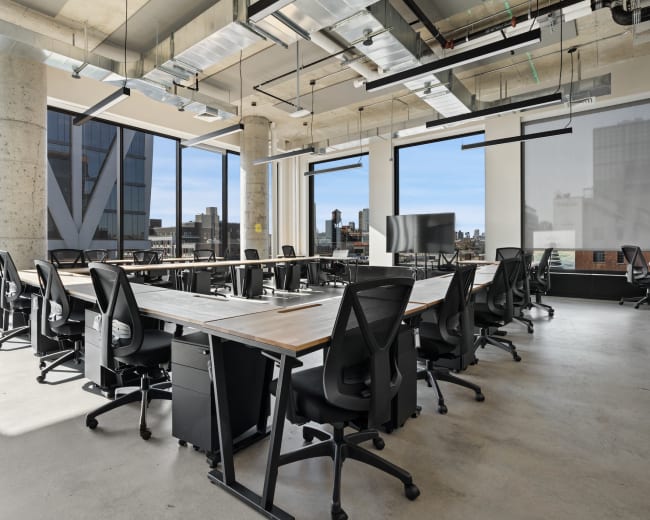 The image shows a modern office space with several long tables and ergonomic chairs arranged neatly, surrounded by large windows offering a view of the city.