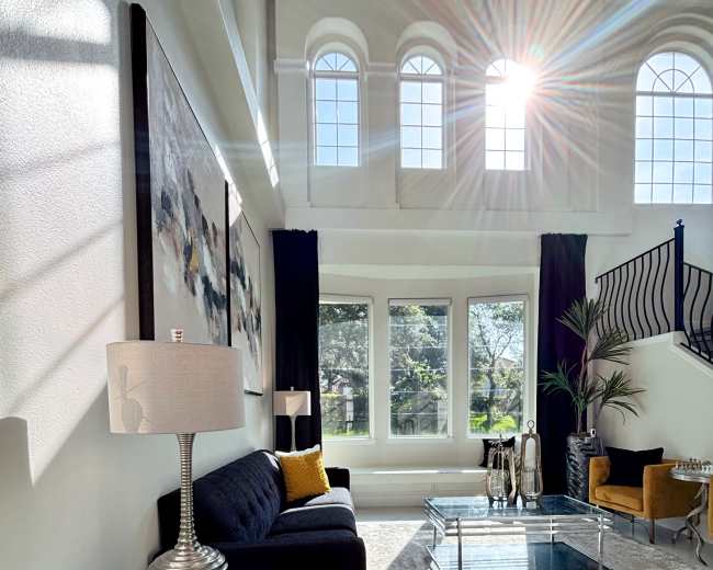 A modern living room features a dark sofa, two glass coffee tables, and large windows allowing sunlight to stream in, illuminating the space.