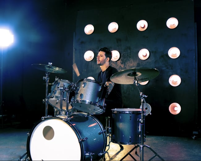 A drummer plays a drum set in front of a wall with circular light fixtures.