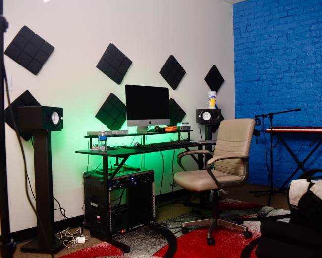 The image shows a small music production studio featuring a desk with a computer, two studio monitors, a keyboard, and a comfortable chair against a backdrop of blue and white walls.