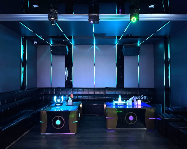 The image shows a dimly lit karaoke room with black leather seating, illuminated by blue and green lights.