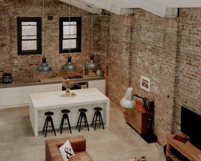 The image shows a modern kitchen and living area within a loft, featuring exposed brick walls, a central island with seating, and industrial-style lighting.