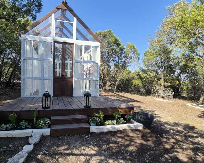A glass-fronted greenhouse with wooden accents is situated among greenery, featuring a wooden deck, lanterns, and landscaped flower beds.