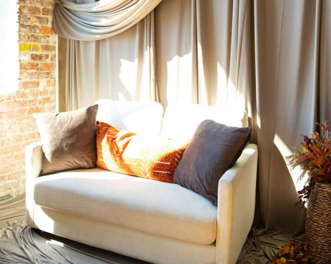 A light-colored sofa with decorative pillows sits against a backdrop of draped beige fabric and a brick wall, complemented by autumn-themed decor.