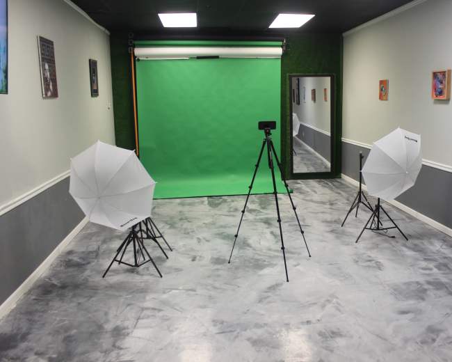 The image shows a photography studio setup with a green backdrop, two softbox lights on tripods, and a camera on a tripod in a sparsely decorated room.