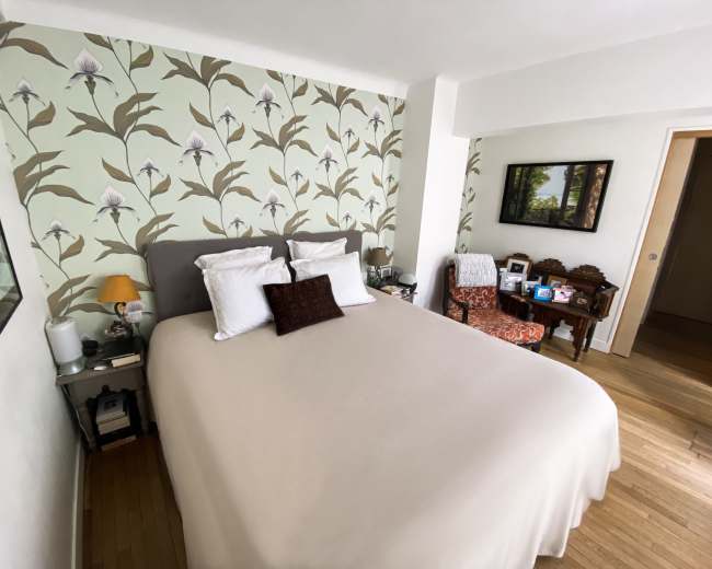 The image shows a bedroom with a patterned wall, a bed with white linens, and a small seating area with an armchair and a table next to it.