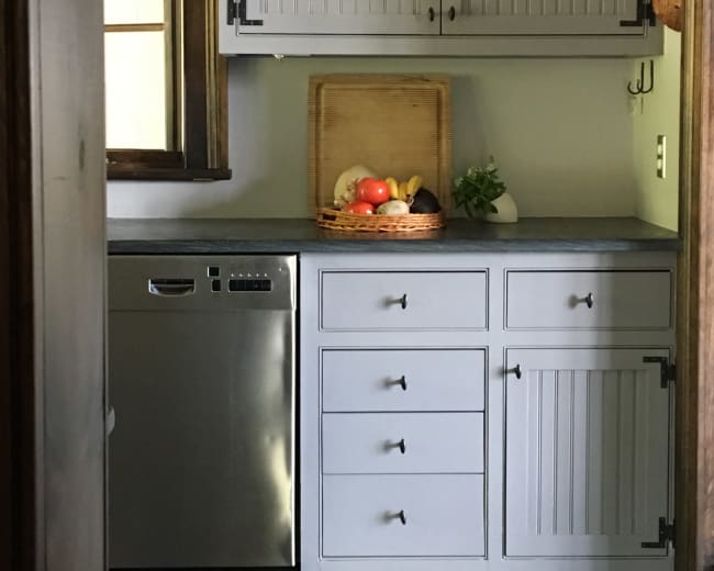 A kitchen features gray cabinets, a stainless steel dishwasher, and a fruit basket on the countertop.