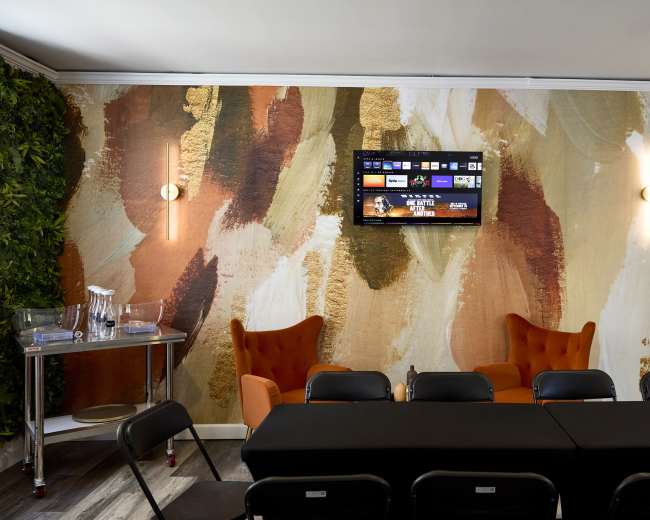 The image shows a modern interior space with two orange armchairs, a long black table, and a wall decorated with large abstract patterns in earthy tones, complemented by a television mounted above.