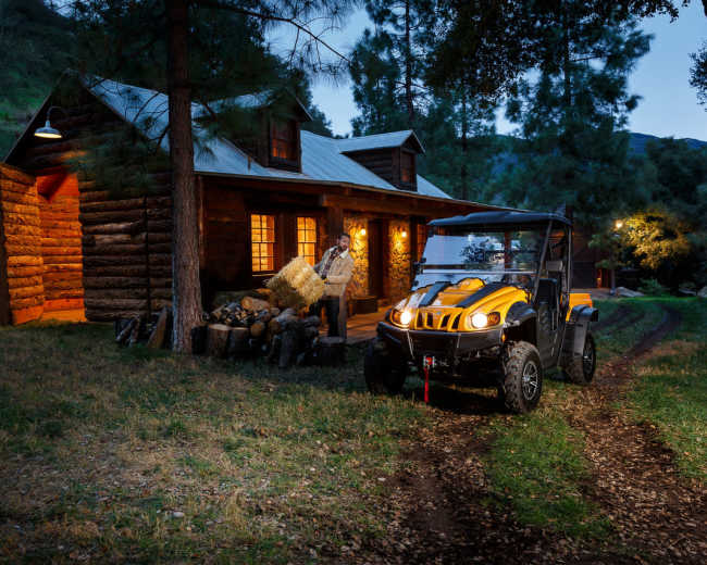 A yellow utility vehicle is parked near a log cabin surrounded by trees, with a pile of firewood and a winding dirt path leading up to the cabin.