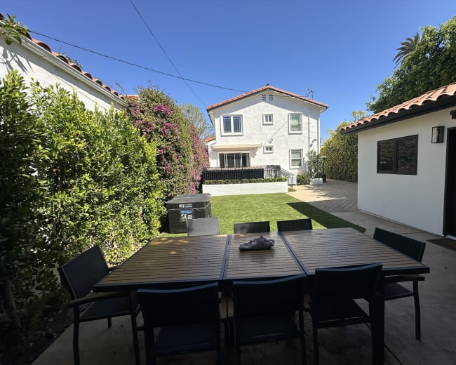 The image shows a backyard patio with a wooden dining table surrounded by black chairs, leading to a house with a garden and a lawn.