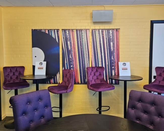 The image shows a seating area with purple bar stools and a round table against a yellow brick wall, featuring two art pieces with colorful vertical lines and a vinyl record.