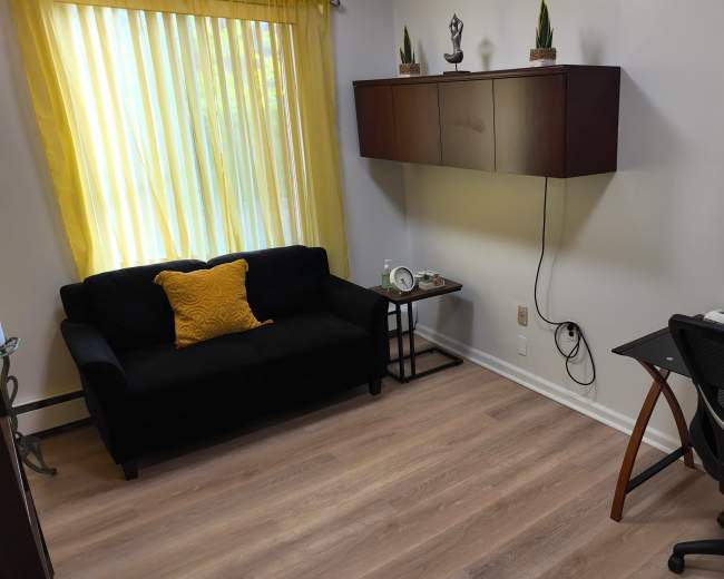 The image shows a small office space with a black couch, a yellow decorative pillow, a desk equipped with stationery, and a wall-mounted cabinet.