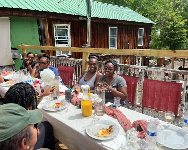 A group of people is gathered around a table on a deck, enjoying food and drinks during a sunny gathering.