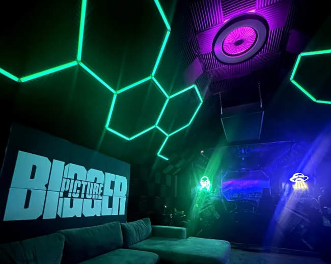 The image shows a modern room with hexagonal neon lights on the walls, a large couch, and a screen displaying the word "BIGGER."