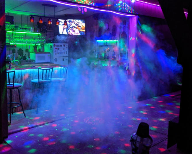 A bar area with illuminated shelves and a television is surrounded by colored lights and a foggy atmosphere.