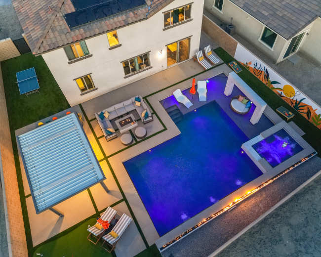 The image shows a modern backyard with a swimming pool, outdoor seating area, and a covered patio, surrounded by landscaping and outdoor decor.