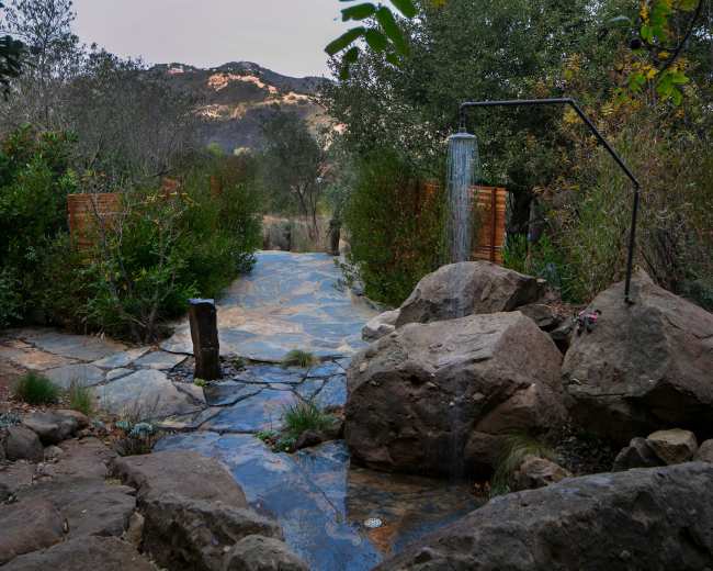 A stone pathway surrounded by greenery leads to an outdoor shower near large rocks and hills in the background.